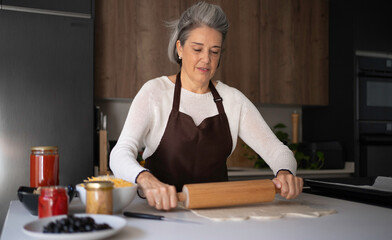 Senior woman wearing an apron rolling dough with a rolling pin in a modern kitchen