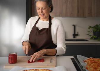 Senior woman wearing an apron, chopping vegetables on a cutting board, preparing a homemade pizza