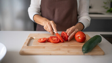 Woman's hands in apron slicing a fresh tomato on wooden cutting board in kitchen