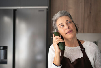 Woman playing with a cucumber, pretending it is a phone in a modern kitchen