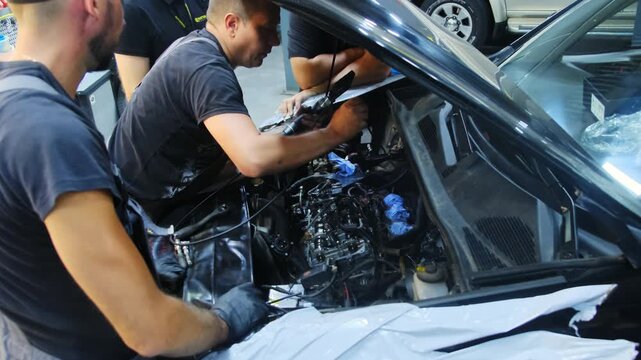 Technician with colleagues checks car using voltmeter in service shop. Mechanics team analyze condition of engine under vehicle open hood