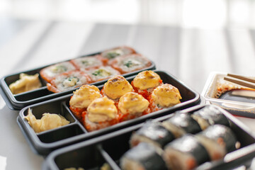 Assorted sushi rolls with baked toppings in plastic delivery boxes served with soy sauce, ginger and chopsticks, concept of Japanese cuisine, food delivery and modern dining at home