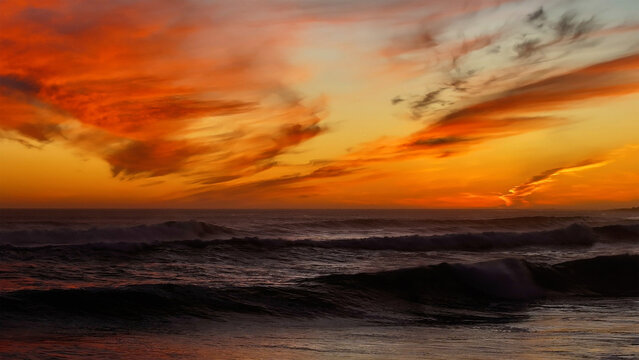 Aerial view of the sun setting over the ocean's dark waves, painting the sky with fiery oranges and yellows, creating a dramatic contrast, Cape Town, Western Cape, South Africa.