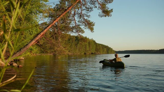 A man in a kayak is paddling across a quiet forest lake at sunset. A traveler in a hat is canoeing near the lakeshore beneath an overhanging tree. A man on a packraft is enjoying the natural scenery