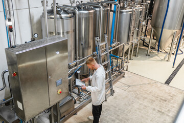 Engineer checking parameters on a tablet, monitoring stainless steel tanks in a beverage production facility