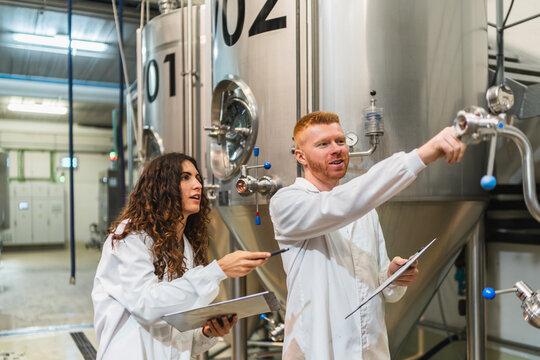 Two workers in lab coats inspecting stainless steel fermentation tanks, discussing quality control processes in a modern brewery