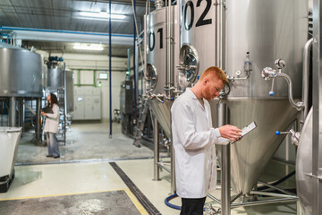 Brewery worker wearing a lab coat, controlling the fermentation process using a digital tablet inside a modern beer factory