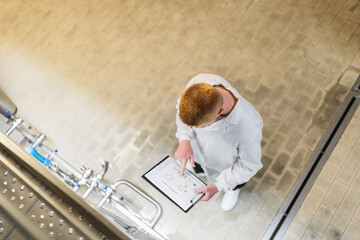 Worker in a white lab coat using a digital tablet for quality control and monitoring beer brewing in an industrial facility