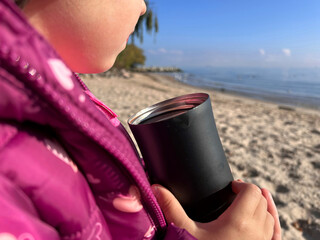 Close-up of a child in a pink jacket holding a black thermos cup on a sandy beach with the sea in the background