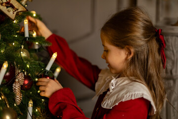 Side view shot of cute little child girl in red fashion dress is decorating Christmas tree in living room at night at home. Christmas Eve and Happy New year winter holidays. High quality photo