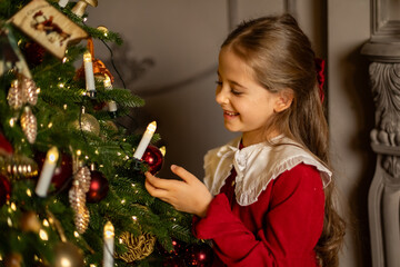 Side view shot of cute little child girl in red fashion dress is decorating Christmas tree in living room at night at home. Christmas Eve and Happy New year winter holidays. High quality photo