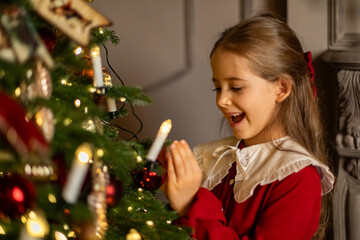 Side view shot of cute little child girl in red fashion dress is decorating Christmas tree in living room at night at home. Christmas Eve and Happy New year winter holidays. High quality photo