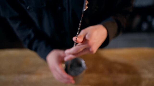 Closeup of a bartender demonstrating barspoon stirring technique above a stainless mixing tin filled with ice, rotating the twisted handle with a firm grip on a wooden counter.