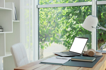 Laptop with empty white screen, lamp, headphones and stationery on desk in room. Comfortable home workplace
