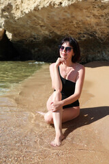 A brunette woman with glasses in a black bathing suit sits on the sand overlooking the sea, with her legs tucked under her, enjoying the sun's rays.