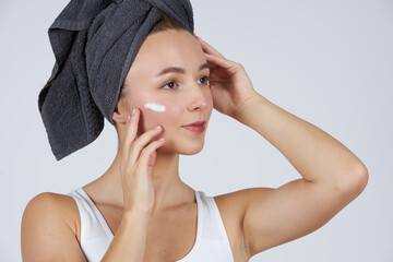 A young beautiful girl in white underwear with a towel on her head uses cosmetic face cream.