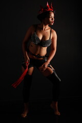 Low key full length portrait of pretty young dark haired woman in leather lingerie with whip in her...