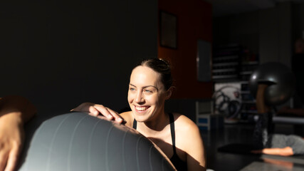 A young woman, smiling, dressed in sportswear, rests during a Pilates class in a small gym