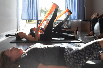 Several women of different ages perform Pilates exercises and stretches, some with elastic bands, in a small gym