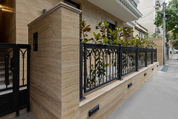 Elegant residential building entrance with beige travertine stone wall, black wrought iron fence...