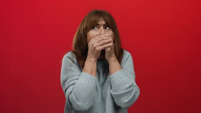 Hispanic senior woman with wide eyes covers mouth in surprise against a bold red wall wearing a gray sweater evoking emotions of shock and disbelief in a stark contrast setting.