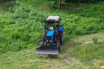 Obraz premium A tractor with an industrial lawn mower is mowing a weed-overgrown lawn. Professional lawn mower. The tractor removes overgrown green grass.