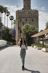 Fototapeta premium A young, beautiful Latin woman with tattoos on her arms and dressed in casual clothes walks and poses on a large avenue in the city on a sunny day. In the background is the golden tower of Seville.