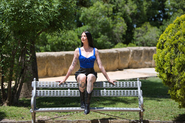 A young, attractive Spanish woman with dark hair, tattoos, a blue top, shorts, and black boots is sitting on a park bench, relaxing in the sun on a spring day.