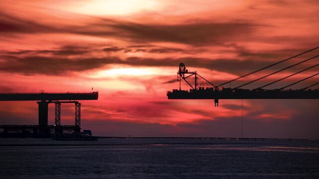 Vibrant sunset with sun partially obscured by clouds over the Tamkang Bridge construction site in Tamsui New Taipei Taiwan. Boat passes under the red and orange sky.