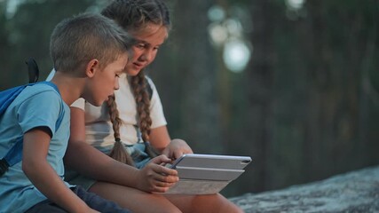 Children using tablet sitting in forest. Boy and girl explore screen content in outdoor nature. Friends interact with technology. Tablet helps children learn. Outdoor moment of learning in forest. - Powered by Adobe