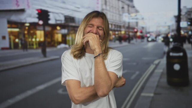 Blond man with long hair and hand on chin laughs in busy street amid blurred pedestrians and glowing storefront lights; amusement fun humor enjoyment.
