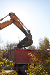 excavator with a large bucket is lifting soil into a red dump truck at a construction site during the day. Clear blue sky is visible above the work area, and trees surround the site