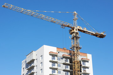 tall construction crane stands beside a multi-story building under development. crane is actively lifting materials against a bright, cloudless sky during daytime