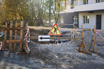 construction site is blocked off by wooden barriers and warning signs. Gravel covers the area with caution tape around the perimeter. setting is a residential street during the afternoon