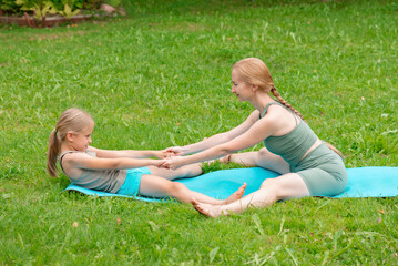 Mother and daughter practice yoga on a mat, stretching outdoors in the summer.