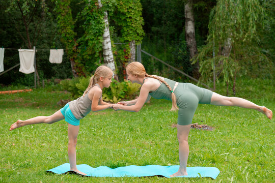 Mother and daughter practice yoga on a mat outdoors in the summer in a country house courtyard.