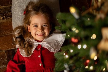 Top view happy smiling little girl with long curly hair in red dress lying under Christmas tree in dark living room at home. Christmas Eve and Happy New year winter holidays. High quality photo