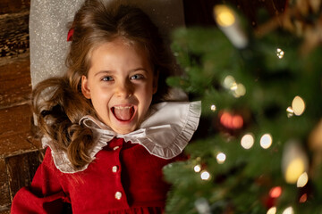 Top view happy laughing little girl with long curly hair in red dress lying under Christmas tree in dark living room at home. Christmas Eve and Happy New year winter holidays. High quality photo