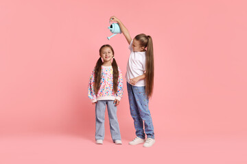 Little girl holding watering can above her friend on pink background. Grow up faster