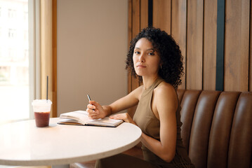 Confident woman sitting at cafe table with open notebook and drink, looking at camera. Concept of freelancer lifestyle, focus, and self development.