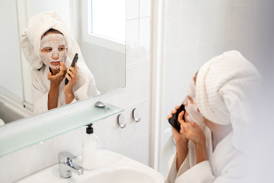 Woman in bathrobe with sheet mask on face talking on smartphone in bathroom near sink and mirror