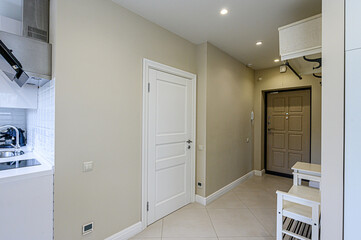 Bright hallway with beige walls, tile flooring, and recessed lighting. White door on the left, brown front door at the end