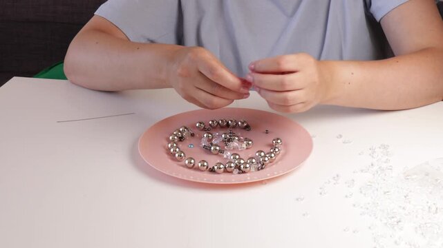 Stringing shiny beads onto a clear fishing line on a pink plate under soft indoor lighting