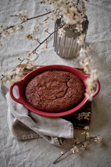 A beautiful and delicious dark chocolate brownie in a burgundy shape on a white tablecloth and a vase with cherry blossoms.