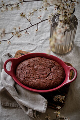 A beautiful and delicious dark chocolate brownie in a burgundy shape on a white tablecloth and a vase with cherry blossoms.