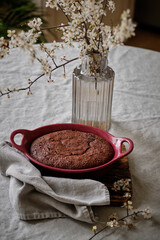 A beautiful and delicious dark chocolate brownie in a burgundy shape on a white tablecloth and a vase with cherry blossoms.