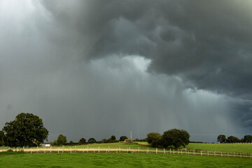 Dark storm clouds rolling across the English countryside, hovering above quiet green fields and wooden fences before a heavy summer rain.