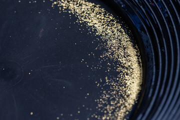 Closeup view of gold flakes collected in a black panning pan. Shiny metallic particles representing mining, prospecting, natural resources, and the search for wealth and treasure.