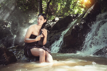 Fototapeta premium Serene Young Woman with Long Hair Sitting by Waterfall in Lush Green Forest, Soft Sunlight Filtering Through Trees, Peaceful Nature Scene