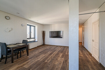 A brightly lit, modern living space with hardwood floors, minimal furniture, and light wallpaper. A dining table is near a window, and a flat-screen TV hangs on the wall